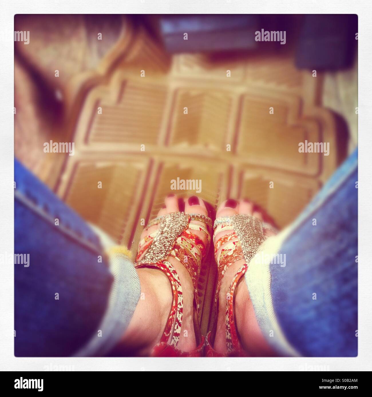 Looking down on blue jeans and painted red toes in ethnic beaded sandals on top of beige plastic Matt in a car. Stock Photo