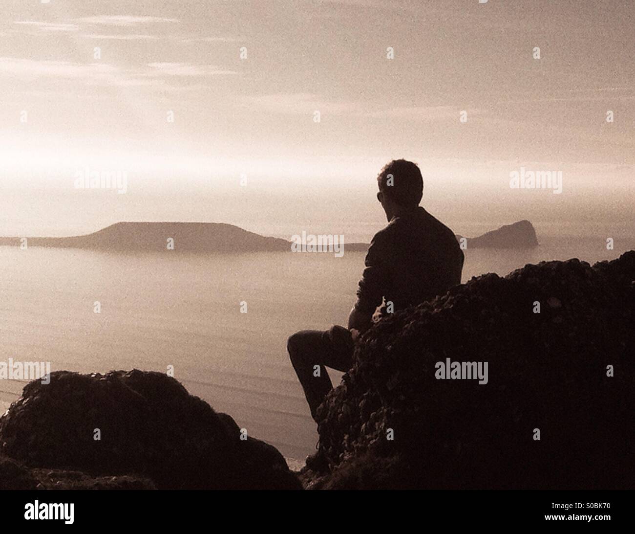 Teenager sitting on a rock overlooking  the sea near Worms head, Gower. Side view silhouette. Stock Photo