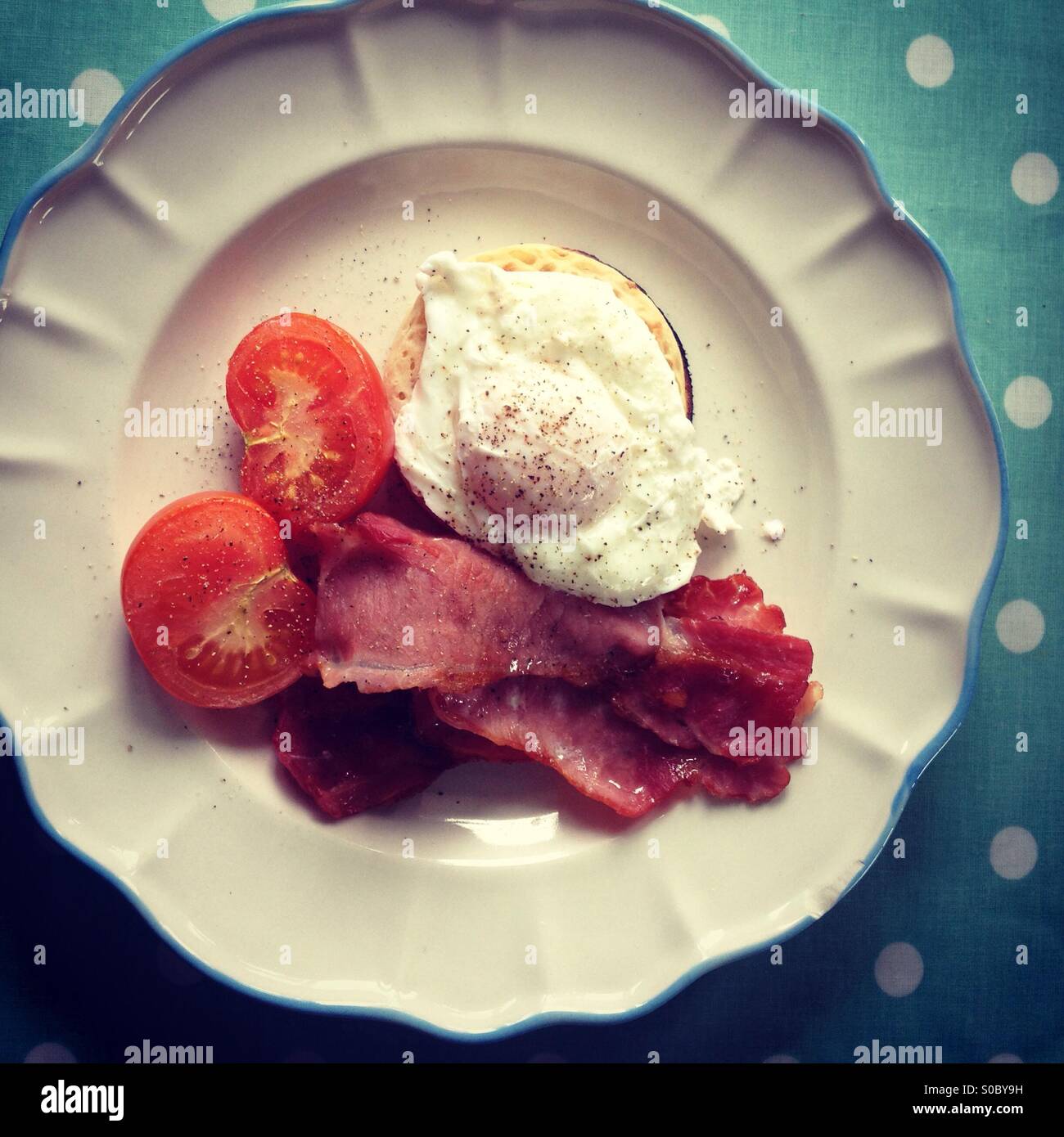 Poached egg on crumpet with grilled tomatoes and back bacon on a white scolloped plate on a blue table cloth with white spots - Smartphone Captured Stock Image