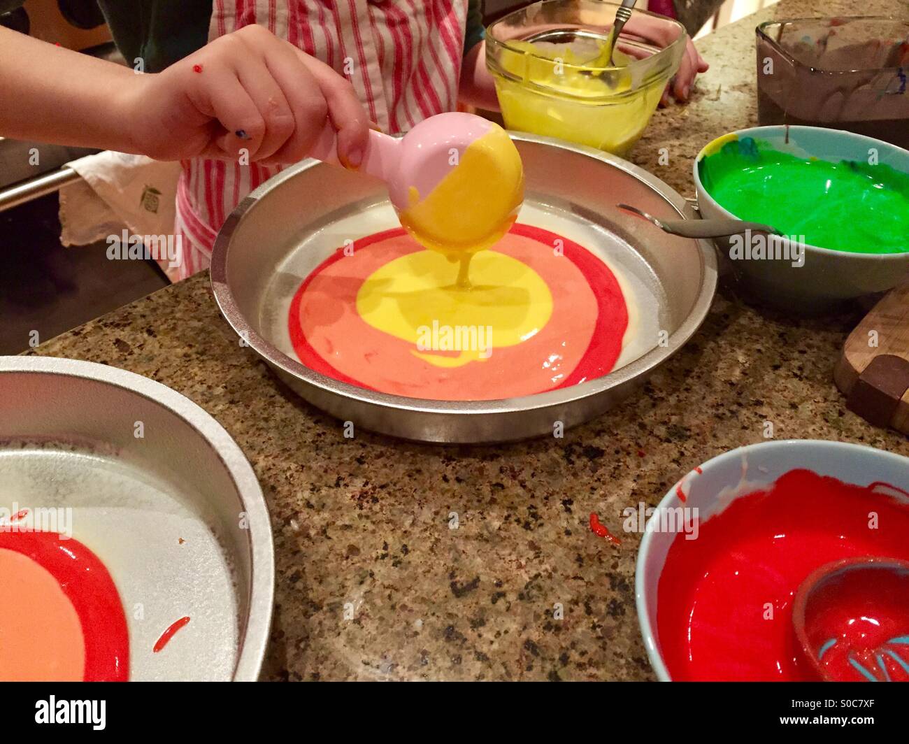 A child pours colorful yellow cake batter into a cake pan on a messy kitchen counter. Stock Photo