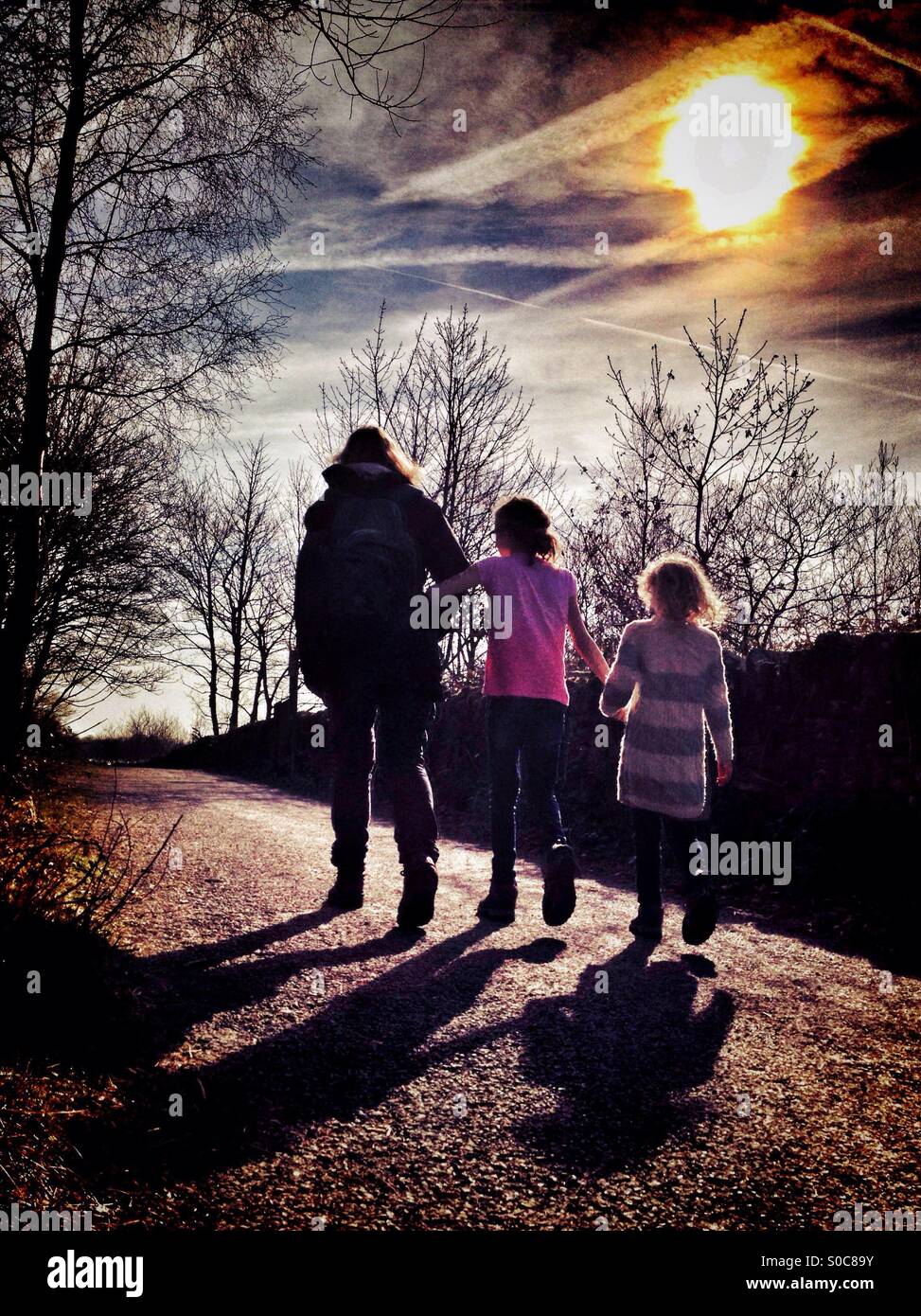Mother and daughters walking holding hands casting shadows and backlit by sun - Smartphone Captured Stock Image