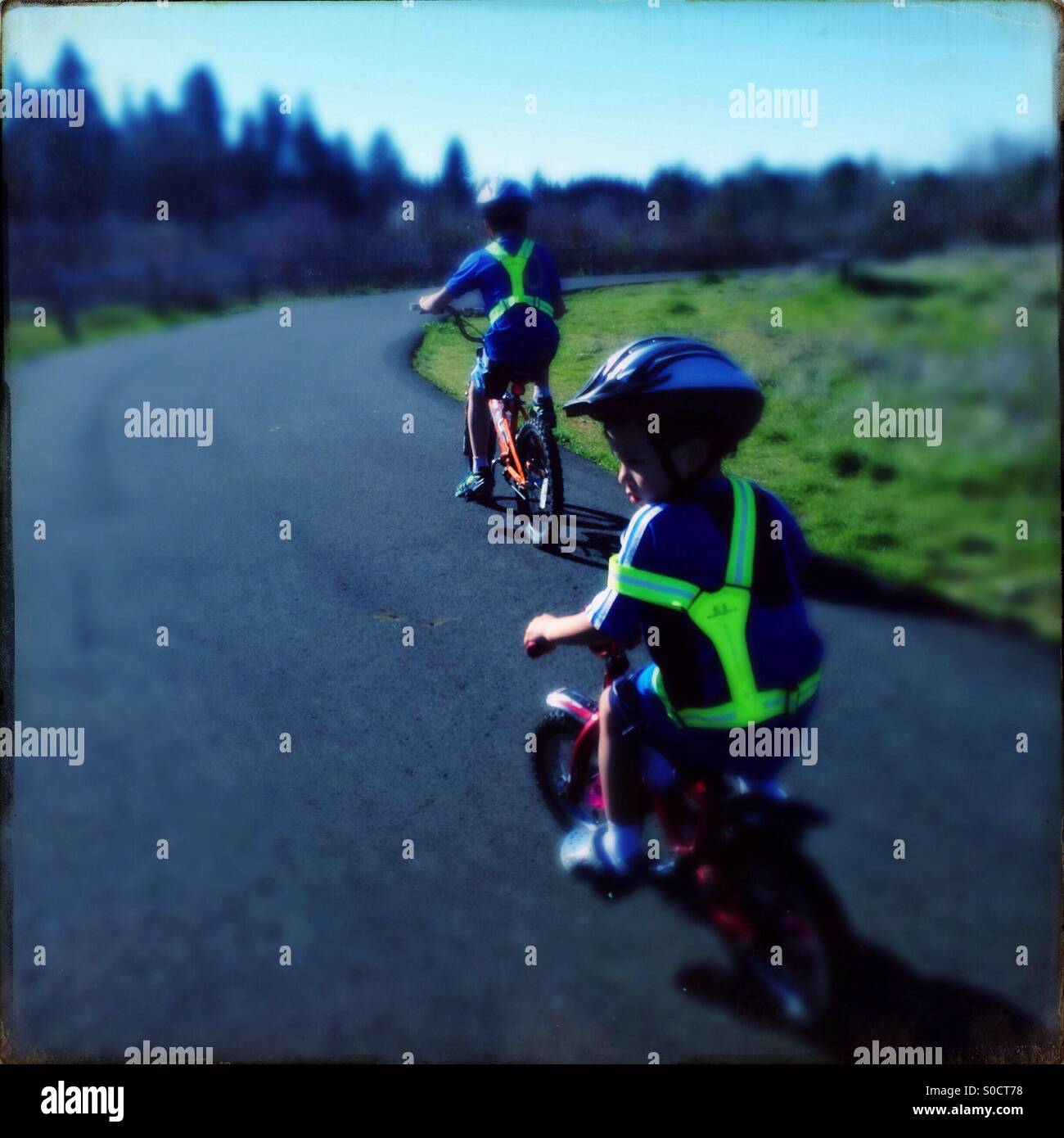 Two boys riding bicycles on path in a park on a sunny day wearing safety vests and helmets Stock Photo