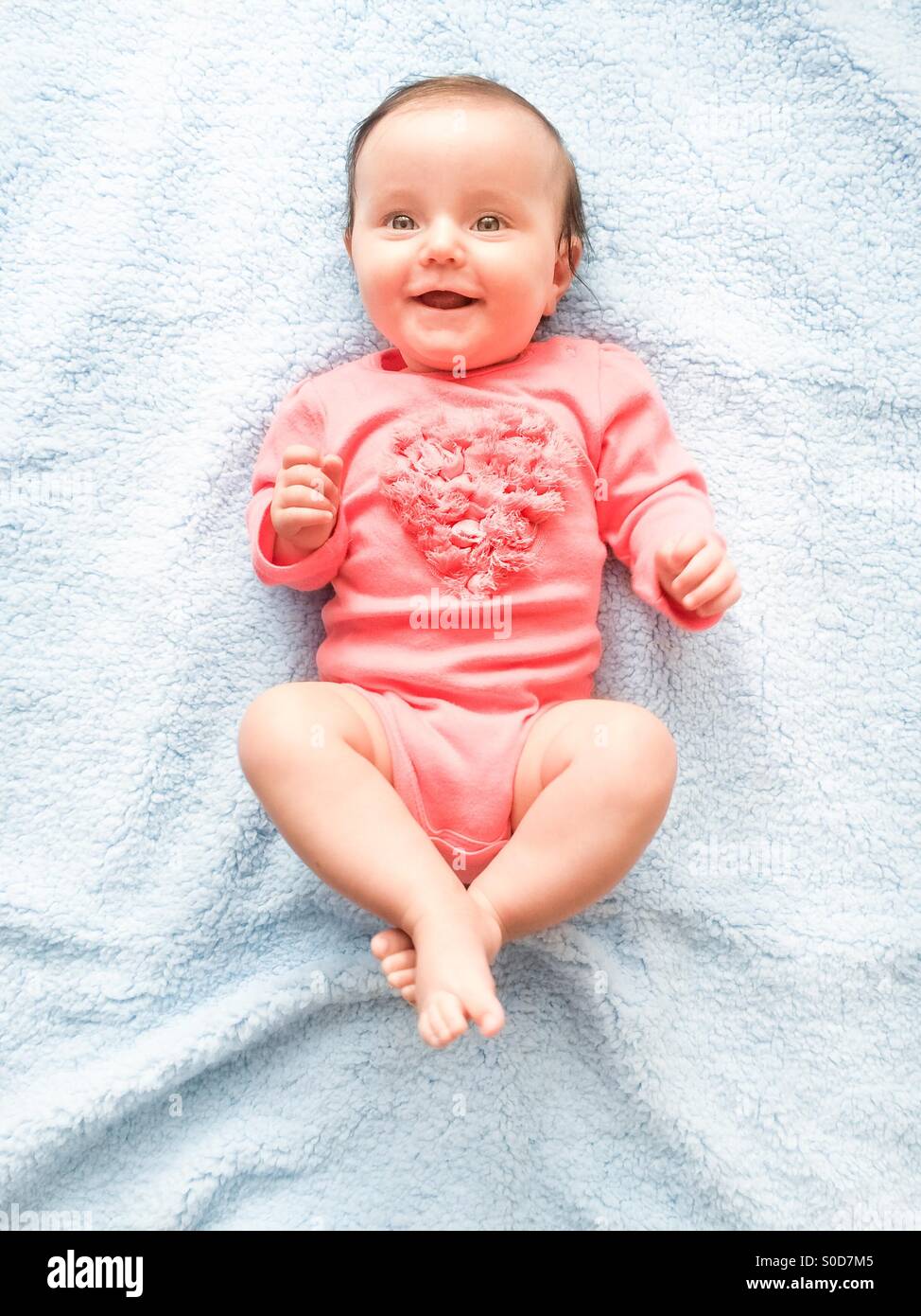 Happy smiling baby girl lying on blue blanket. She is wearing body suit with heart shape. Top shot. Stock Photo