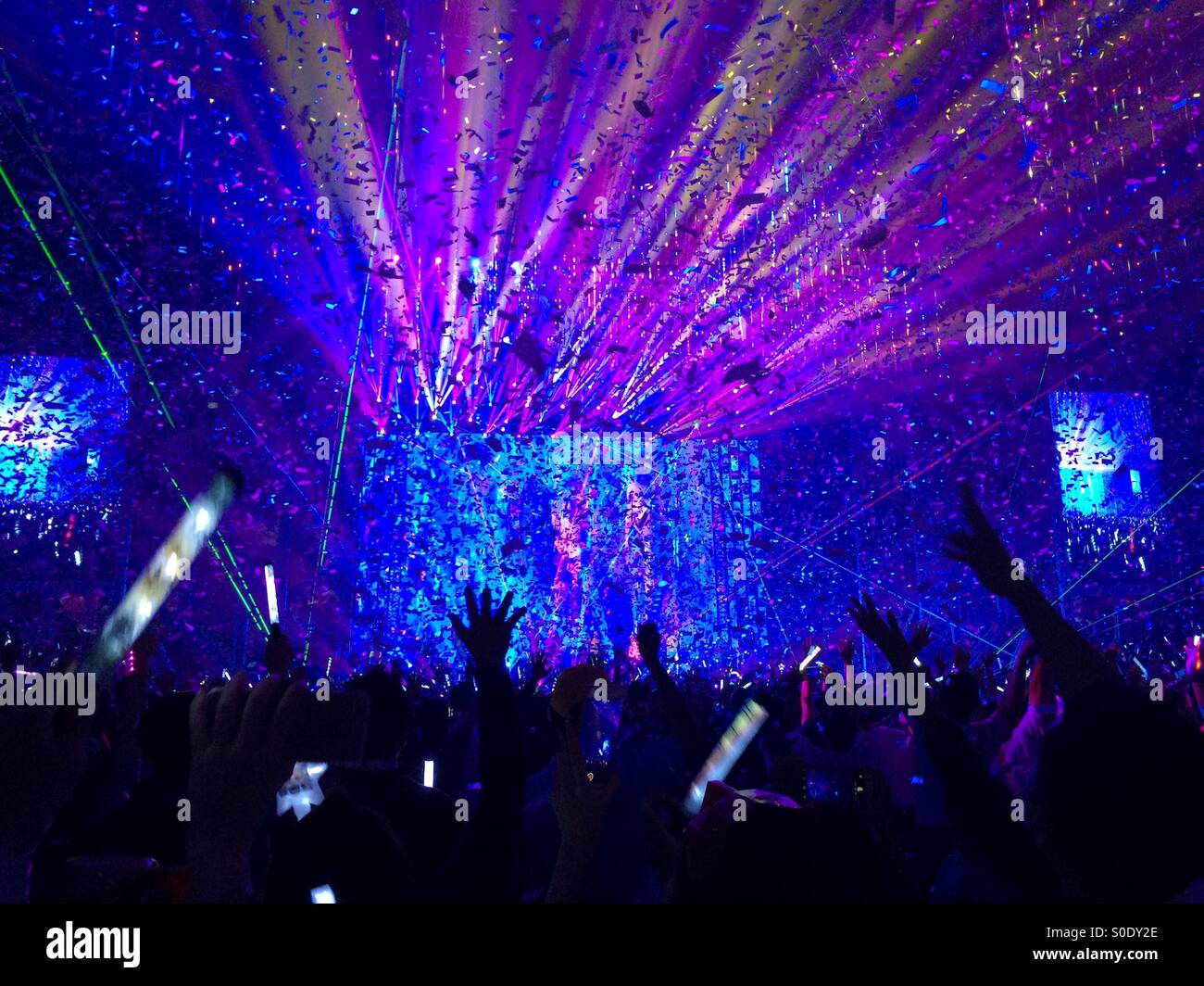 Hands and glow sticks in the air as confetti rain down in a burst of color at the end of a concert. Stock Photo