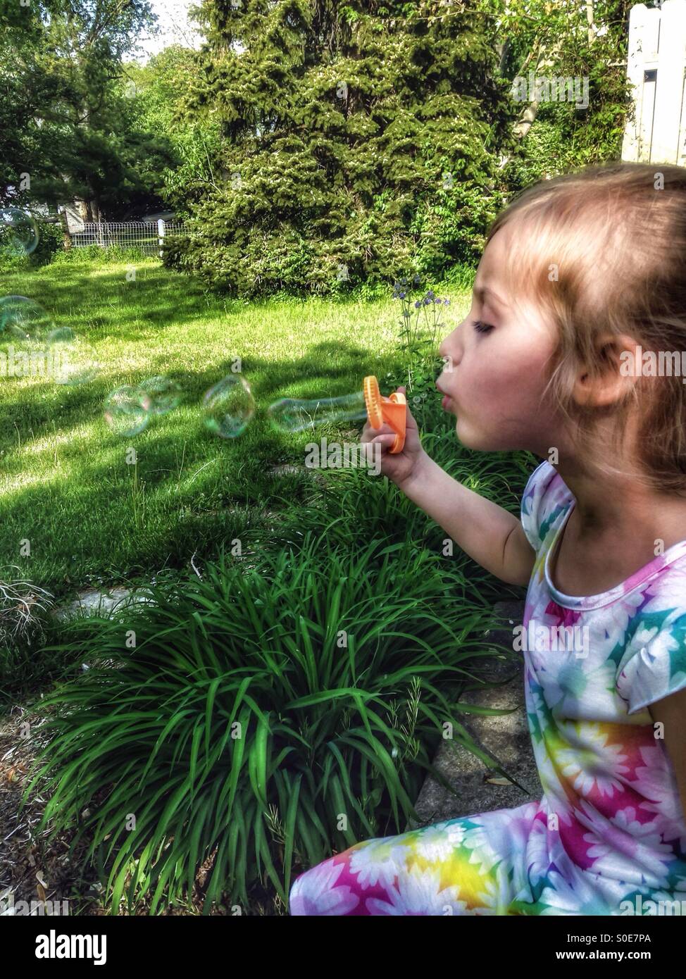 A girl in a floral dress sits on a patio blowing bubbles (2). - Smartphone Captured Stock Image