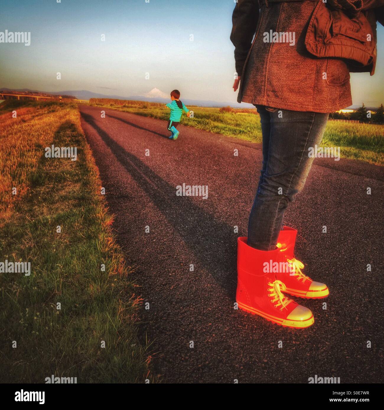 A mom wearing red boots watches while her little boy runs away on the path in the late afternoon. Stock Photo