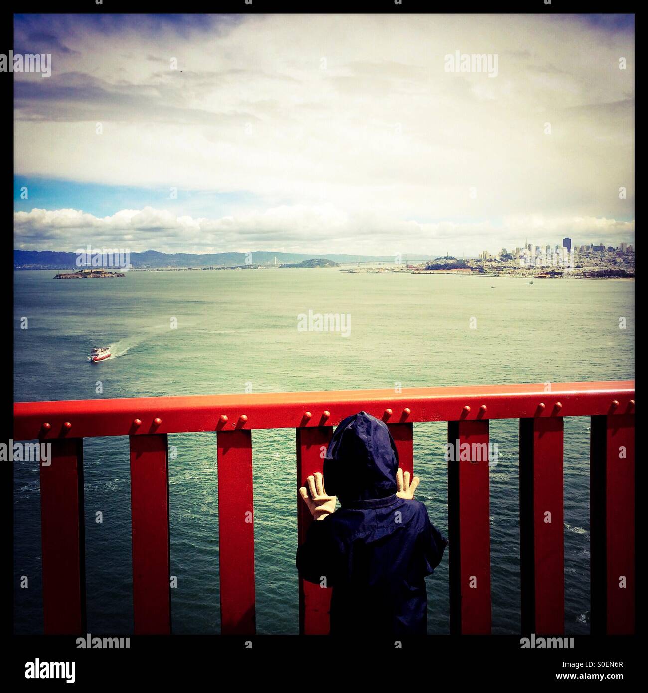A seven year old boy looks at a tourist cruise boat and San Francisco through the rail on the Golden Gate Bridge. San Francisco, California, USA - Smartphone Captured Stock Image
