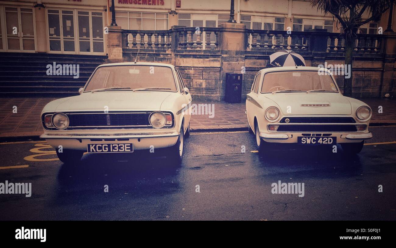 Pair of white Lotus tuned Ford Cortina Mk1 and Mk2 parked on wet seafront road in front of grand architecture at Brighton, Sussex, England. - Smartphone Captured Stock Image