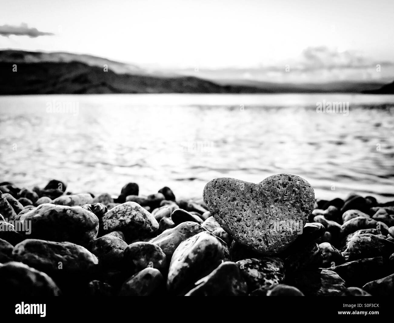 Summer love; in black and white. Heart shaped rock on the beach with lake and mountains in the background. - Smartphone Captured Stock Image