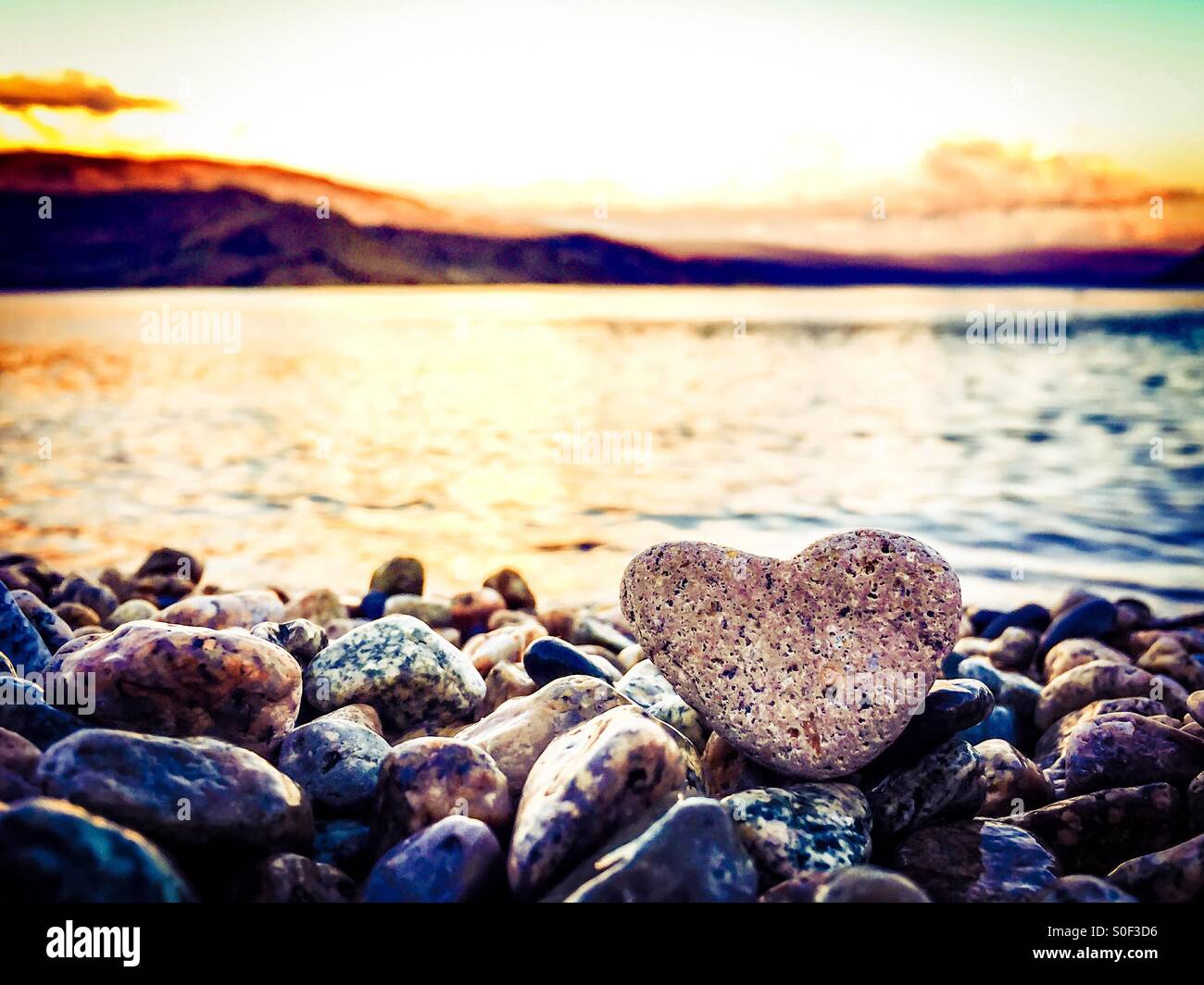 Summer Love. The highlight is on the heart shaped rock sitting on the beach in the foreground. Sunset over lake and mountains in the background. - Smartphone Captured Stock Image