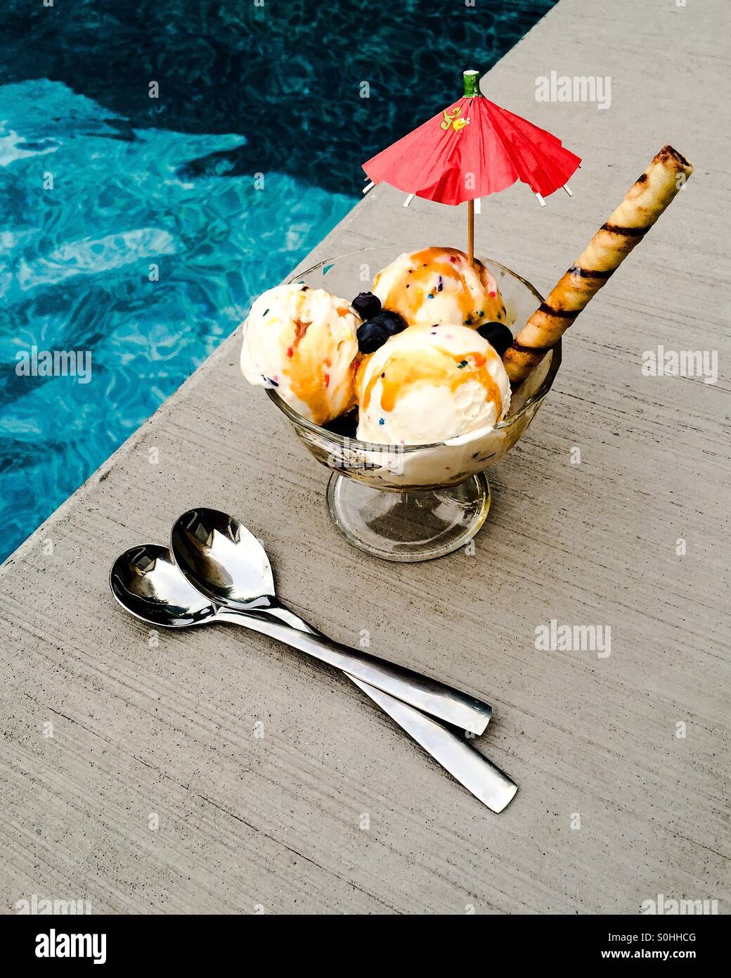 Bowl of ice cream with umbrella and waffle straw in it, sprinkled with blueberries and doused in caramel sauce. There are two spoons next to the frozen treat. Stock Photo