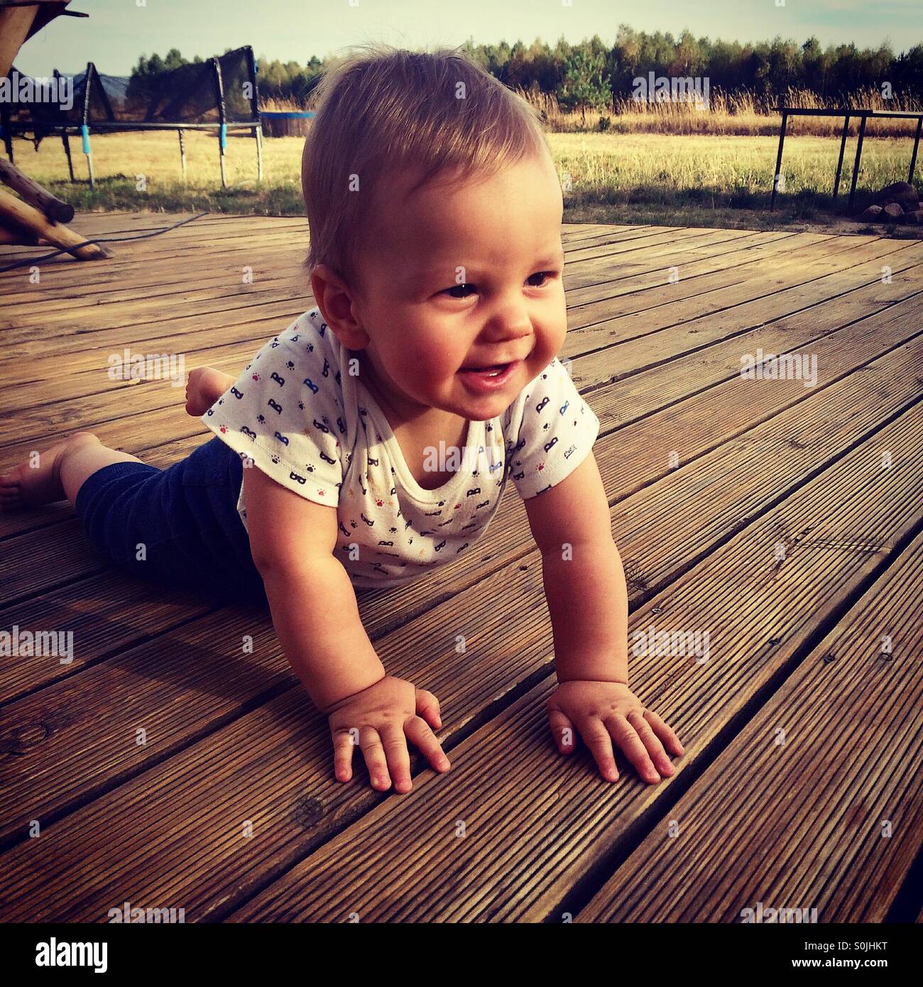 8 months old baby boy learns how to crawl on a wooden deck outside of the house at the back yard on a warm summer afternoon Stock Photo