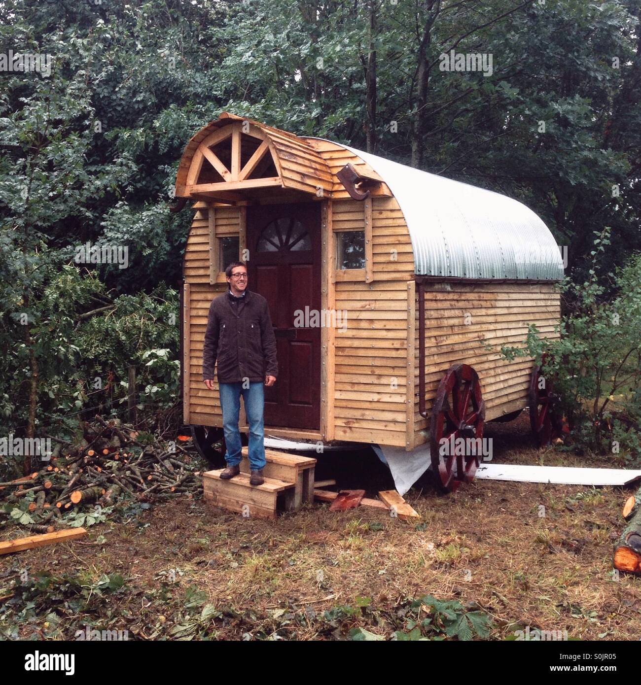 Wooden shepherds hut style log cabin in the woods with a man on the steps, Hampshire, England, United Kingdom. - Smartphone Captured Stock Image