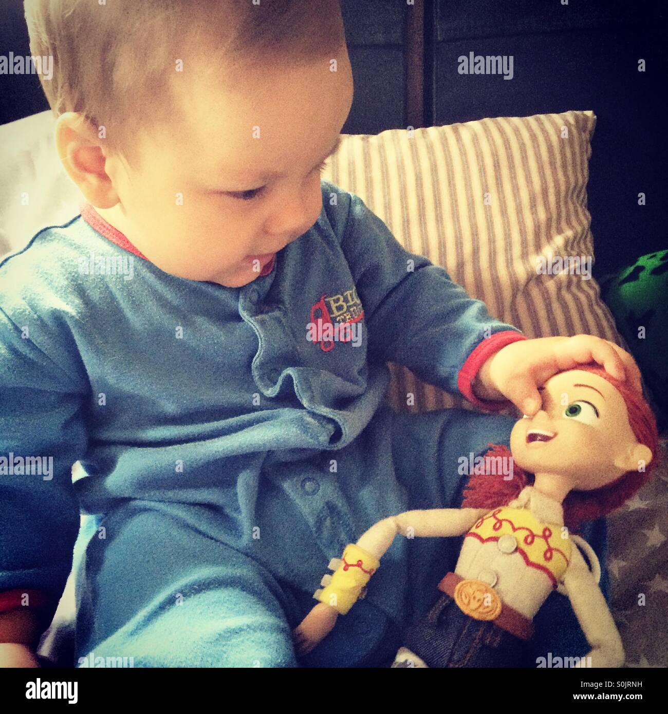 Baby boy wearing blue pajamas is sitting on a blanket with pillows at the back and is playing with his sister doll , a character from Toy Story- Jessie . Stock Photo