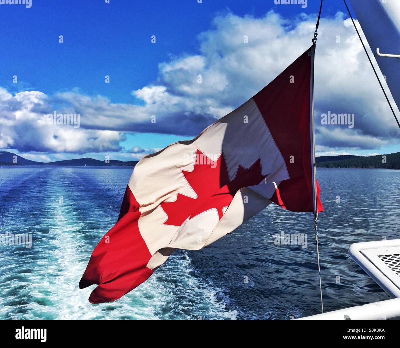 Canadian flag flying in the wind at the back of large boat with the blue sea and sky in the background - Smartphone Captured Stock Image