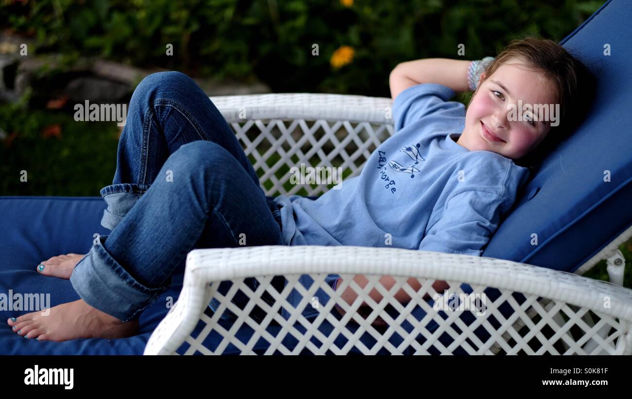 Sweet young girl lounging on chair outdoors wearing jeans and a t-shirt and smiling for the camera. Stock Photo