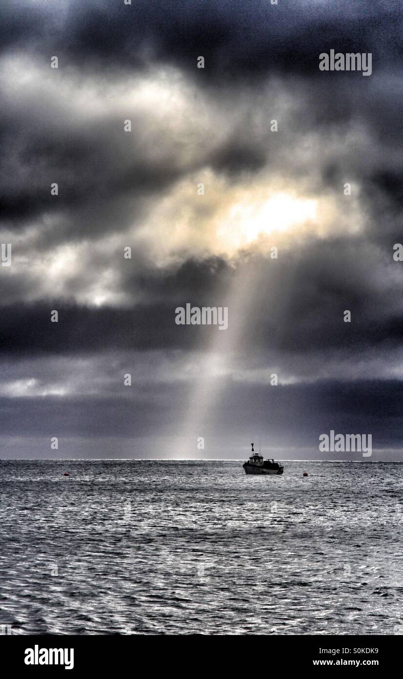 A break in storm clouds lets out a shaft of sunlight over the ocean. A small fishing boat floats nearby. - Smartphone Captured Stock Image