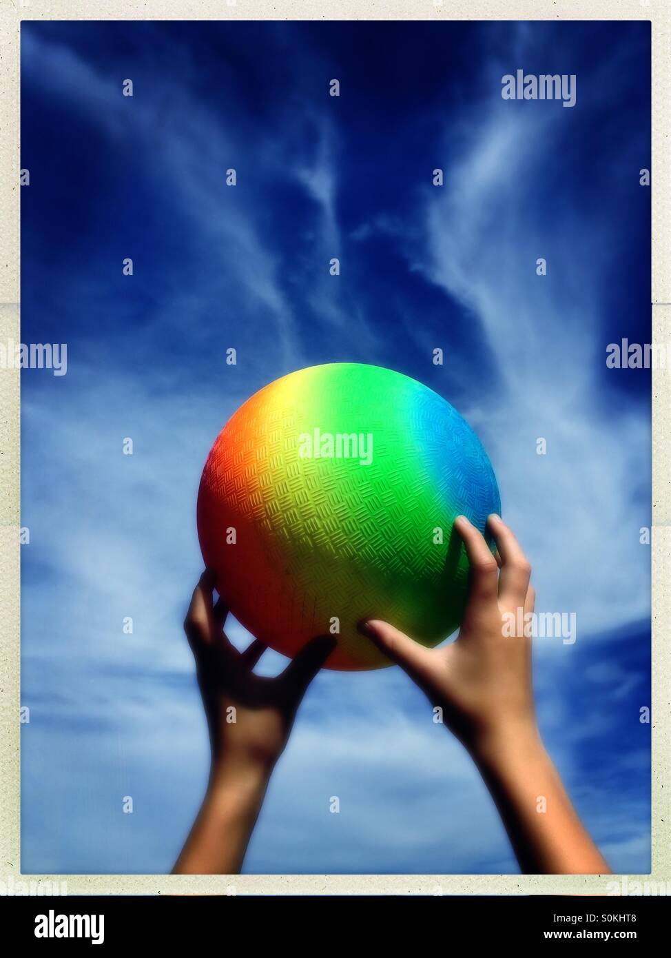 Child reaching for a rainbow colored ball against a deep blue sky. Stock Photo