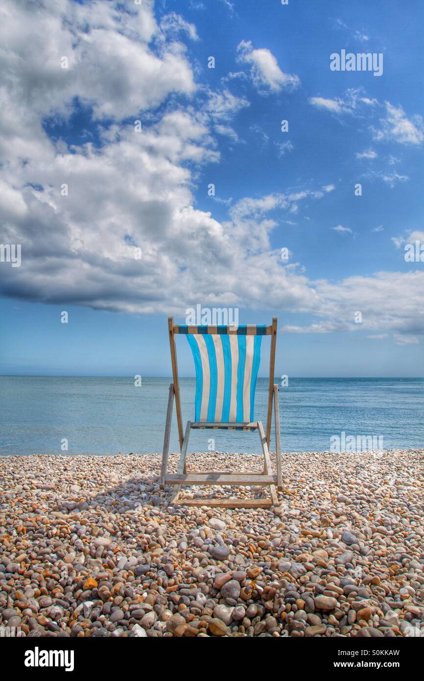 A single blue and white striped Deckchair sits on a deserted pebble beach at the height of summer. - Smartphone Captured Stock Image