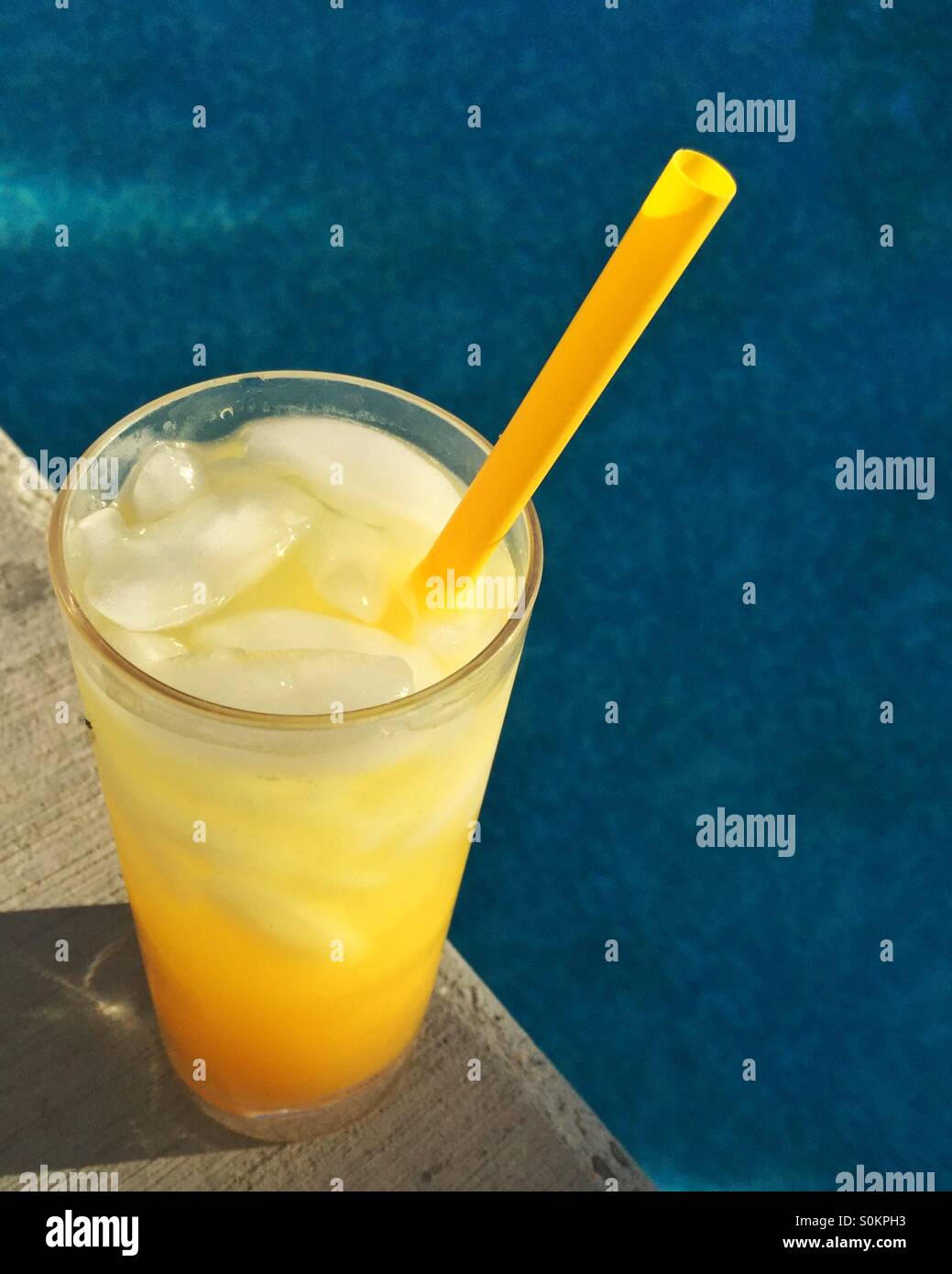 A glass of tropical drink full of ice cubes sits poolside in the afternoon sun. - Smartphone Captured Stock Image