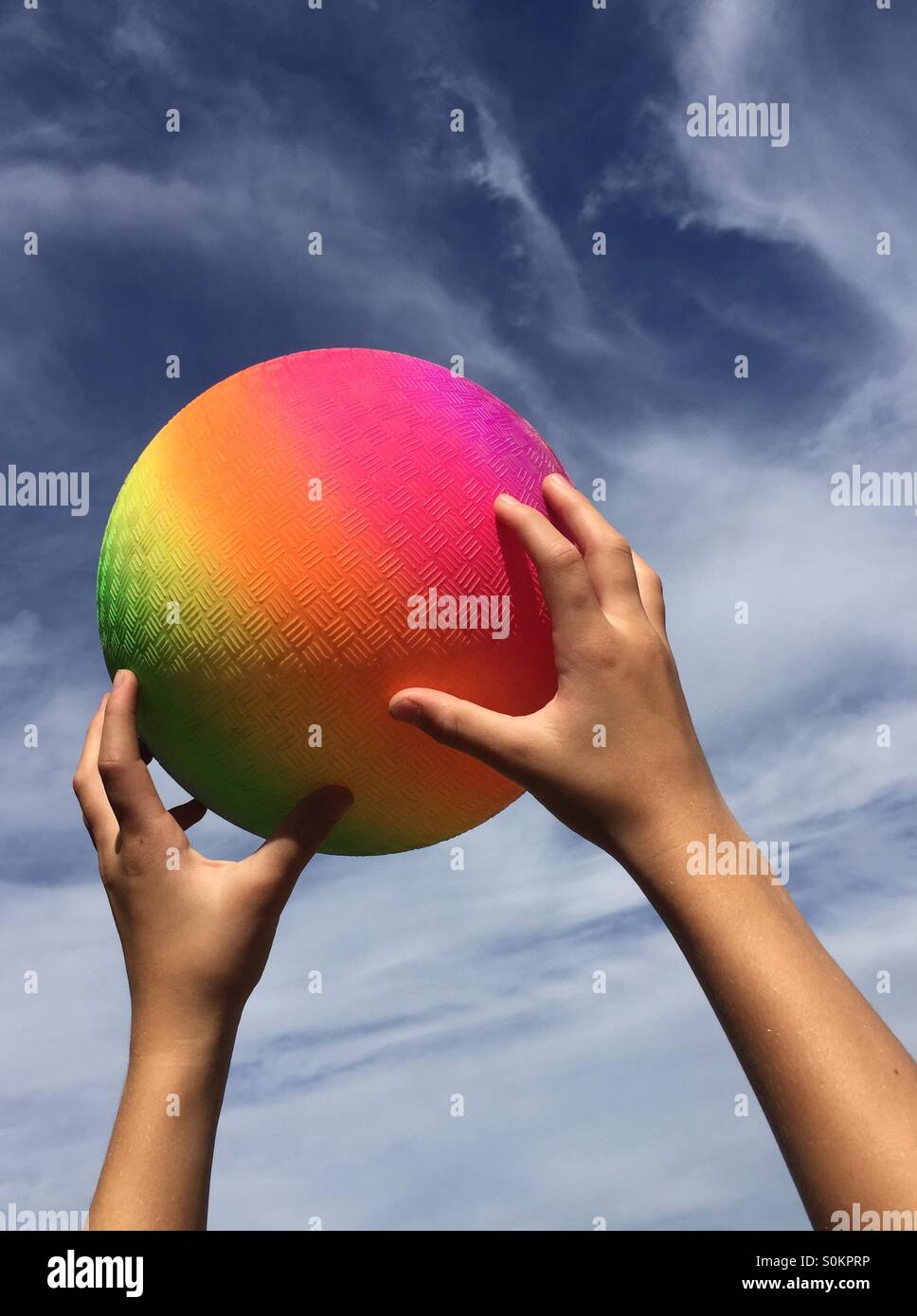 Childs hands catching a rainbow colored rubber ball against a hazy blue sky Stock Photo