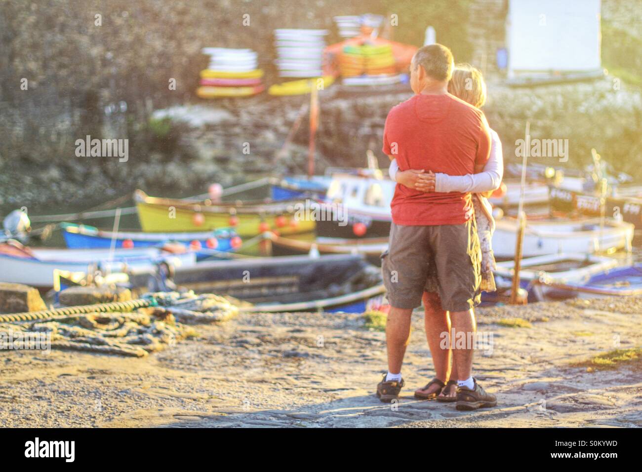 Two people hug on a Cornish harbour at sunset with fishing boats behind them. - Smartphone Captured Stock Image