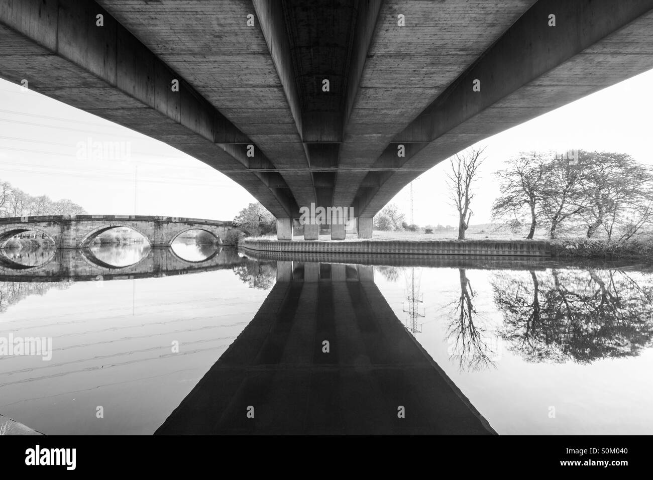 Black and white image of the underside of a concrete bridge with an old bridge further down the river. - Smartphone Captured Stock Image