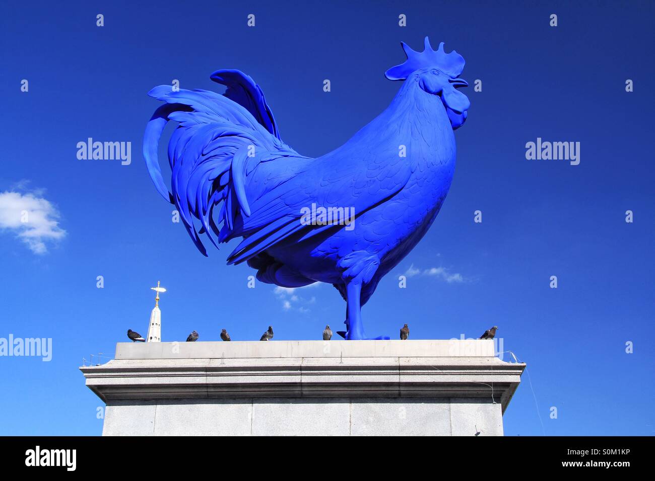 A big, stone, blue chicken statue sits on top of a plinth in Trafalgar Square in London with a blue sky behind it and some pigeons as company. - Smartphone Captured Stock Image