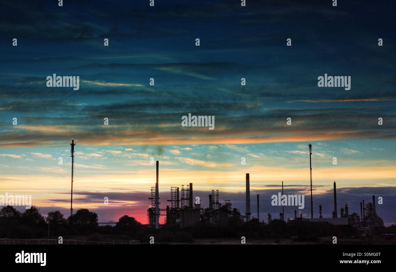 The chimneys of an oil refinery in silhouette pollute a sunlit sky. Sunrise on a colourful sky. - Smartphone Captured Stock Image