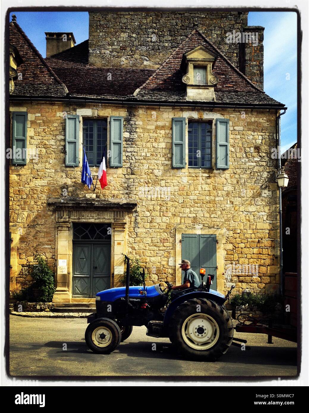 A farmer drives his tractor past the flags on the town hall in rural Domme, Dordogne, France. iPhone photo with some Instagram style processing. - Smartphone Captured Stock Image