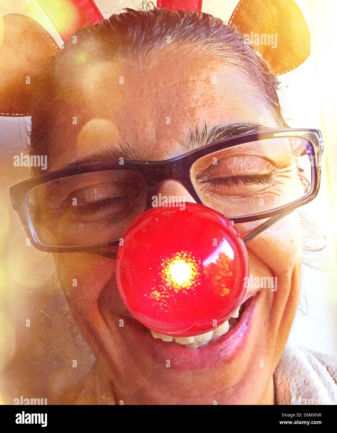 Portrait of a woman laughing and having fun with a blinking red nose and toy reindeer antlers on her head. - Smartphone Captured Stock Image