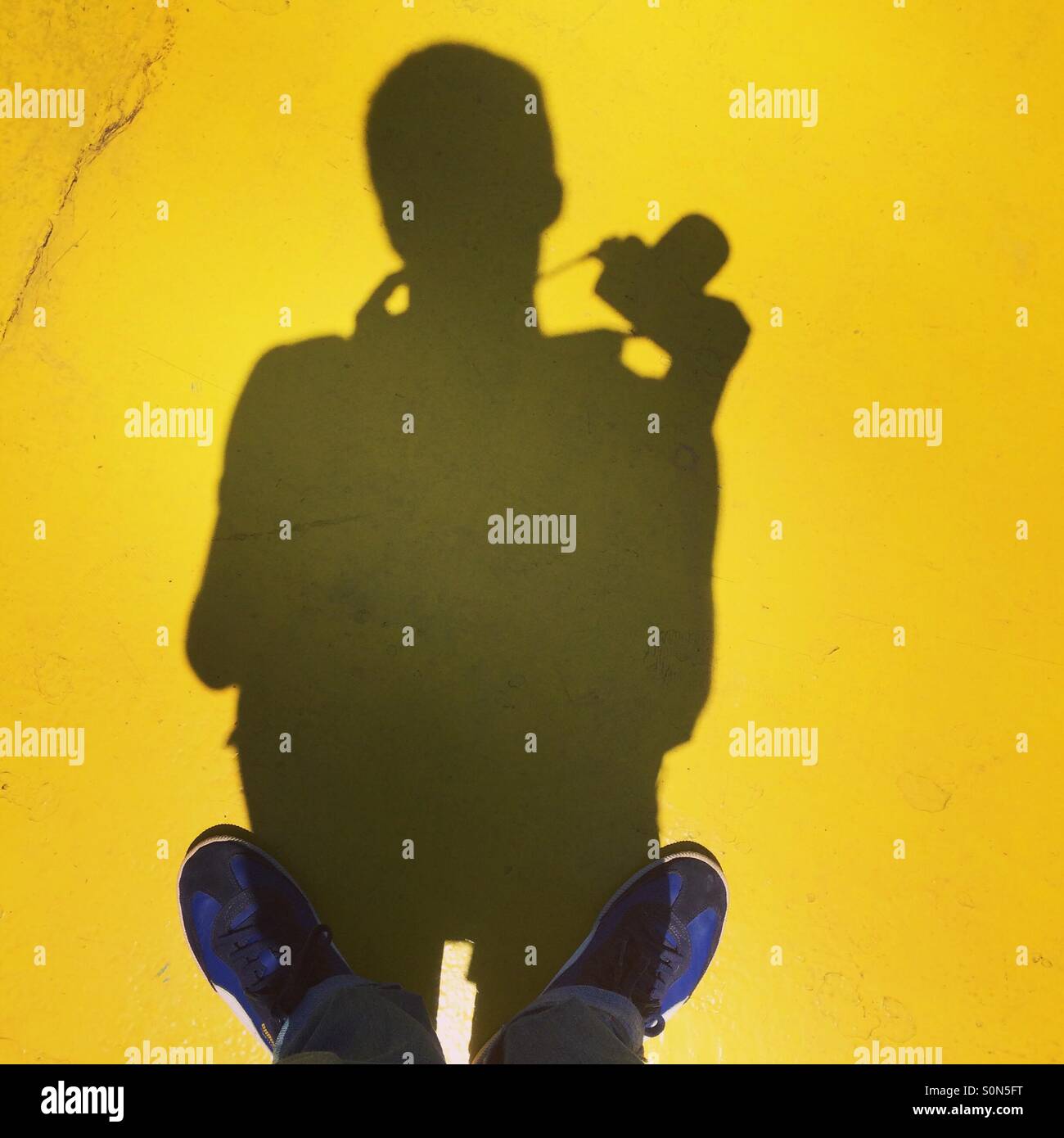 Feet and shadow of a man with a camera against a bright yellow surface. - Smartphone Captured Stock Image