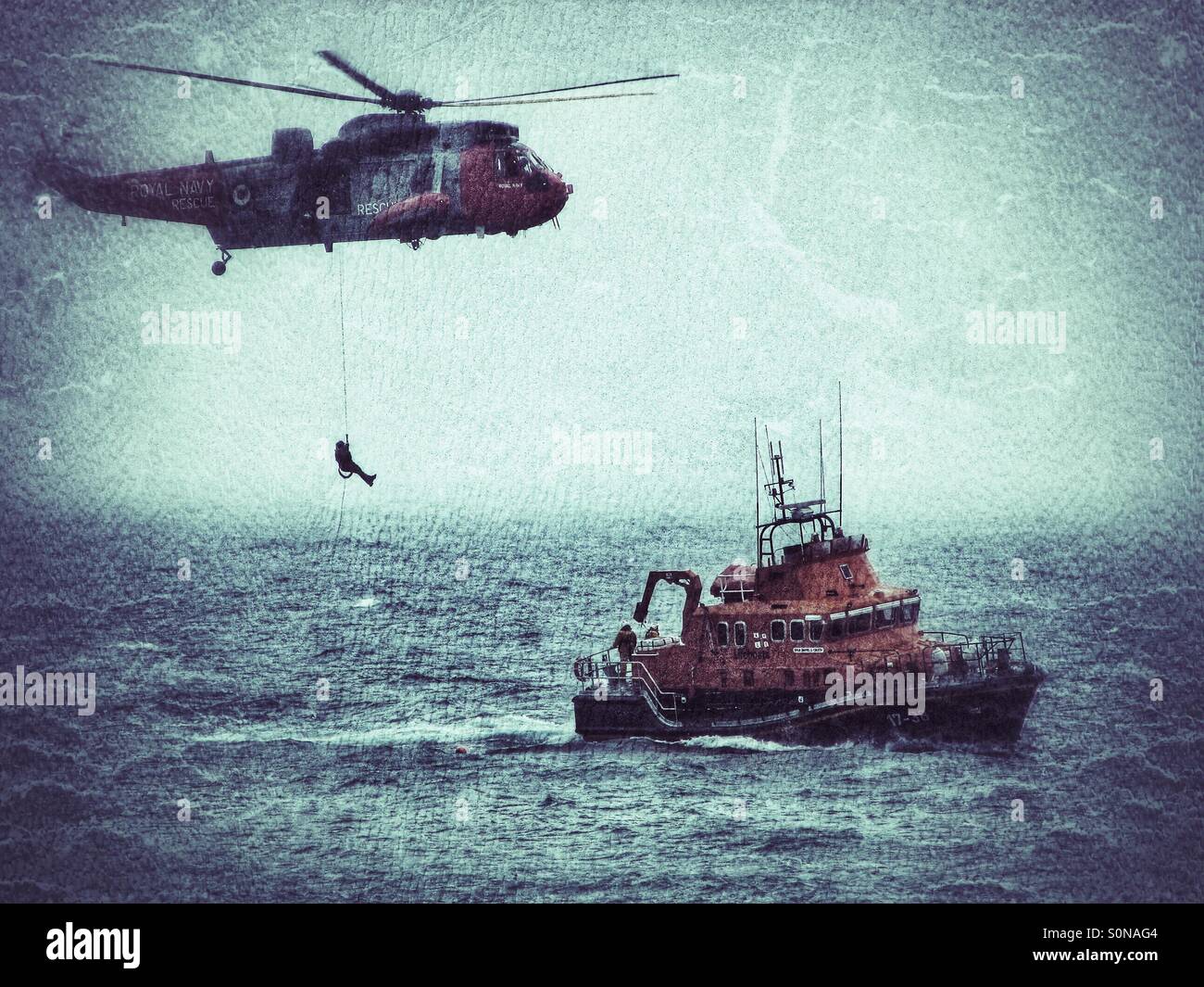 A Royal Navy Merlin Helicopter winches an aircrew member down to a waiting RNLI Lifeboat on a misty sea. Stock Photo