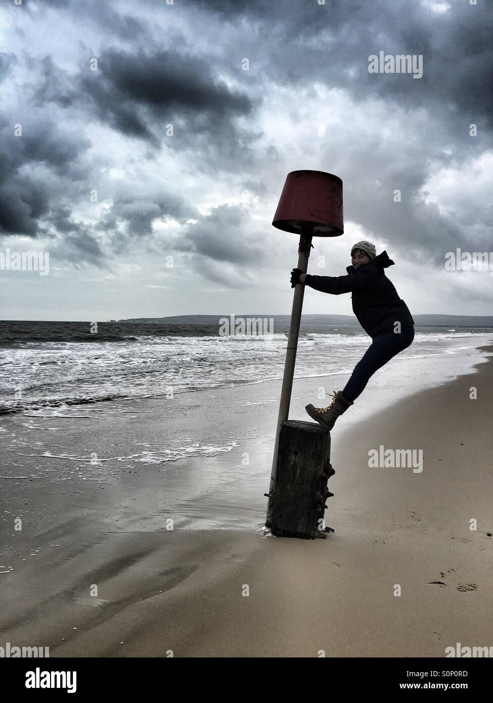 Woman having fun balancing on marker buoy on Dorset beach in winter - Smartphone Captured Stock Image