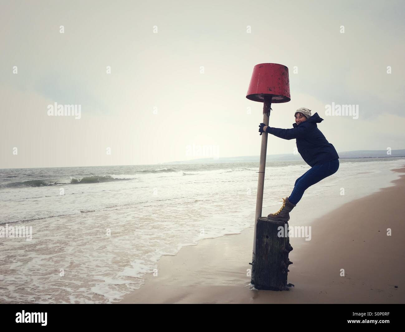 Woman having fun balancing on marker buoy on Dorset beach in winter - Smartphone Captured Stock Image