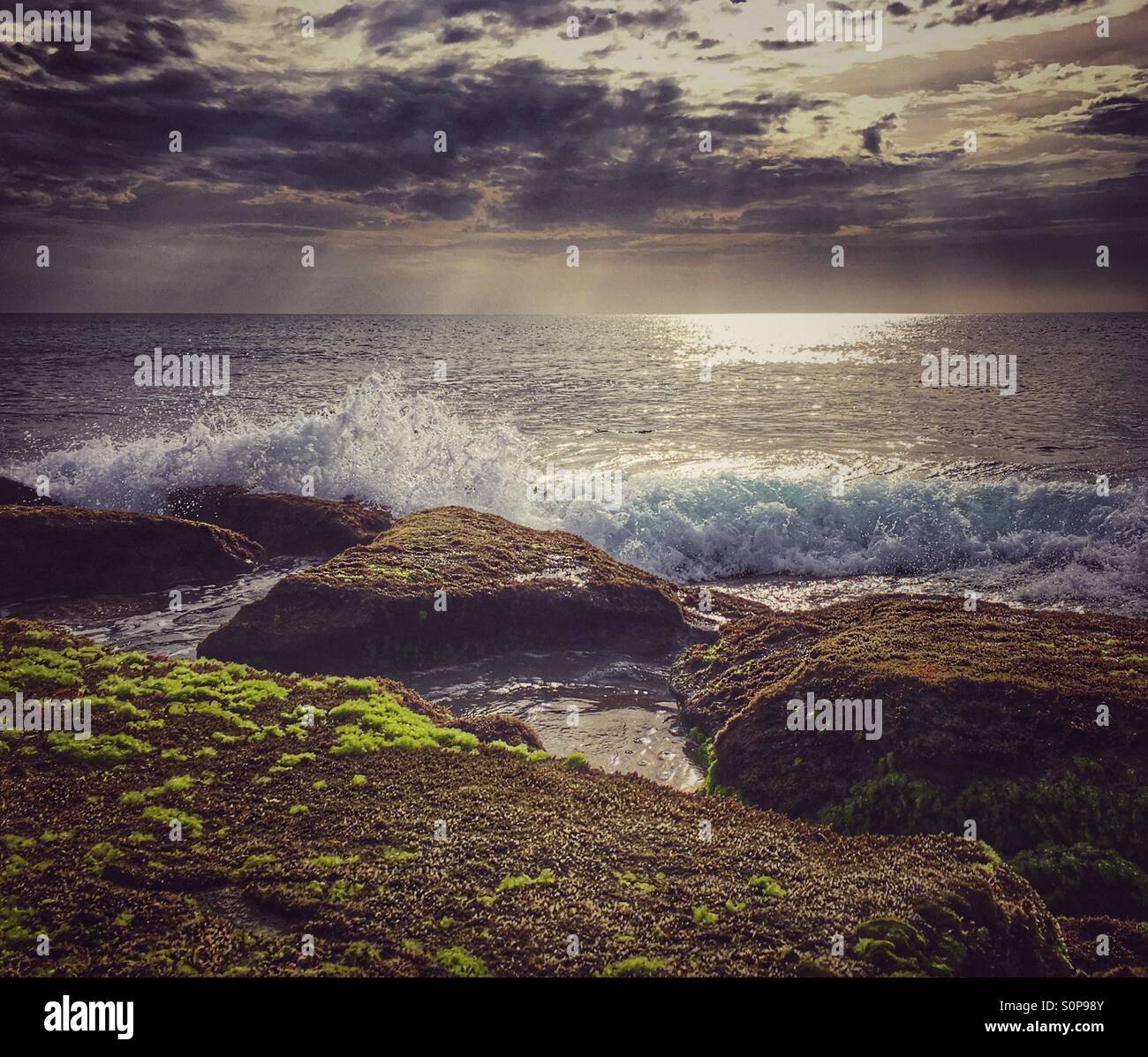 Sunlight streaming through clouds as waves crash into rocks - Smartphone Captured Stock Image