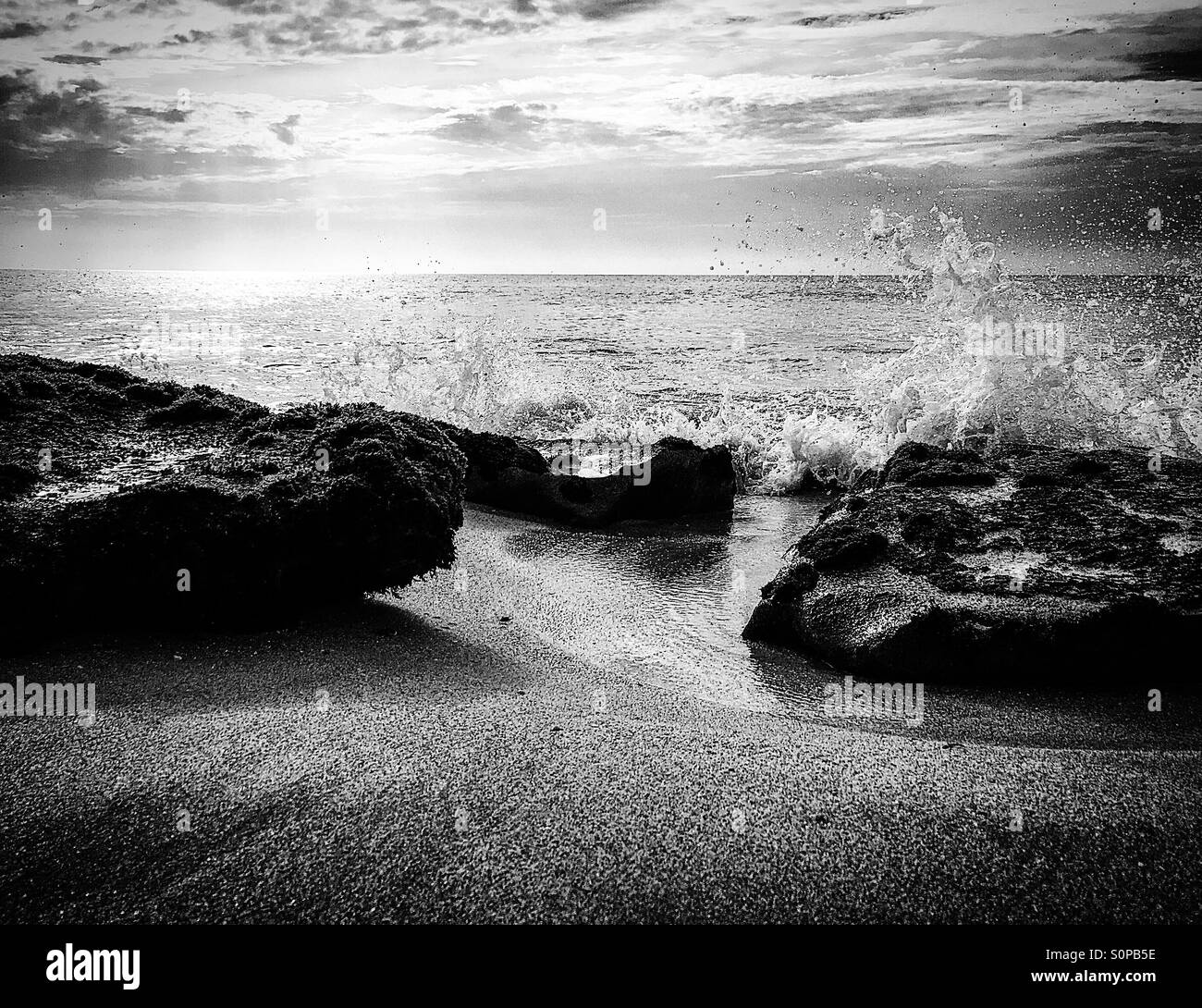 Black and white image of waves breaking over a rock on a sandy beach - Smartphone Captured Stock Image