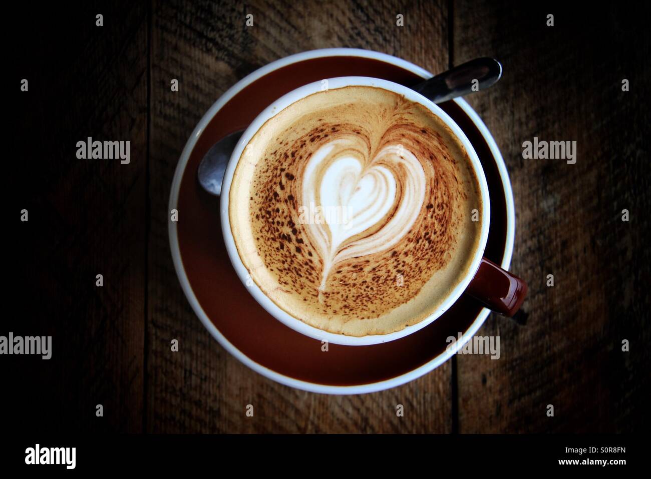 A cup of coffee with a heart shaped pattern sat on a dark table. - Smartphone Captured Stock Image