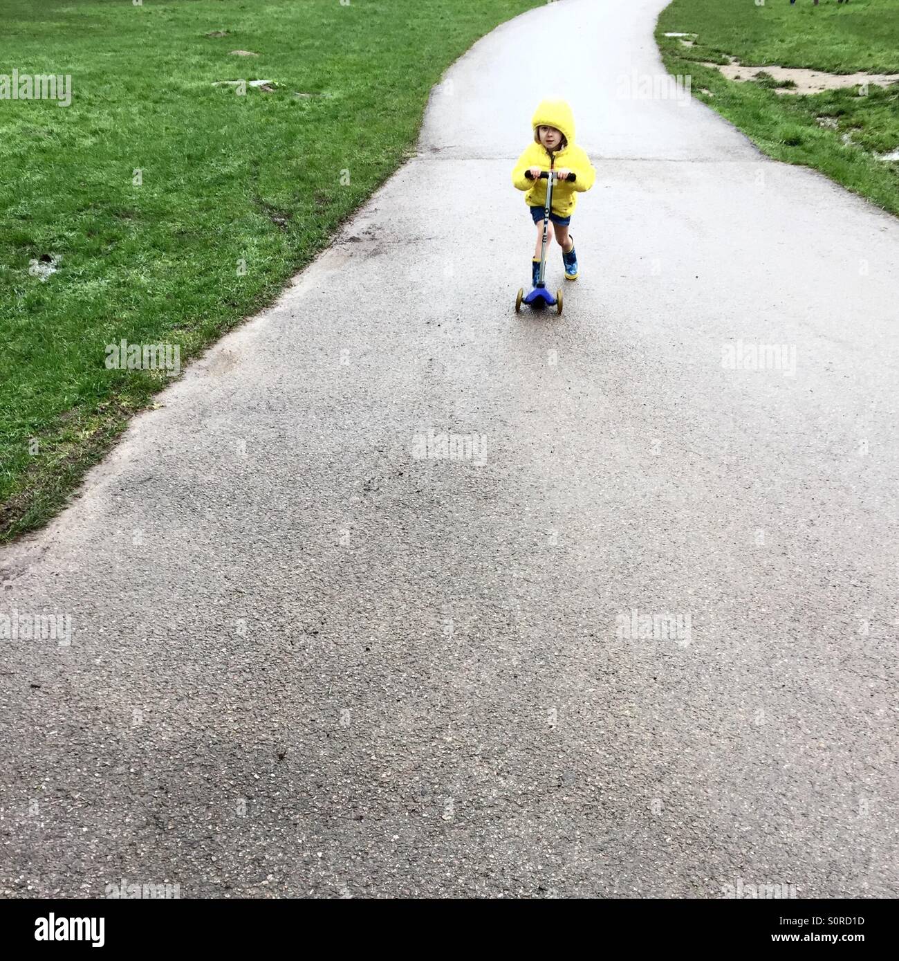 5 year old girl with a yellow coat on a scooter racing Stock Photo