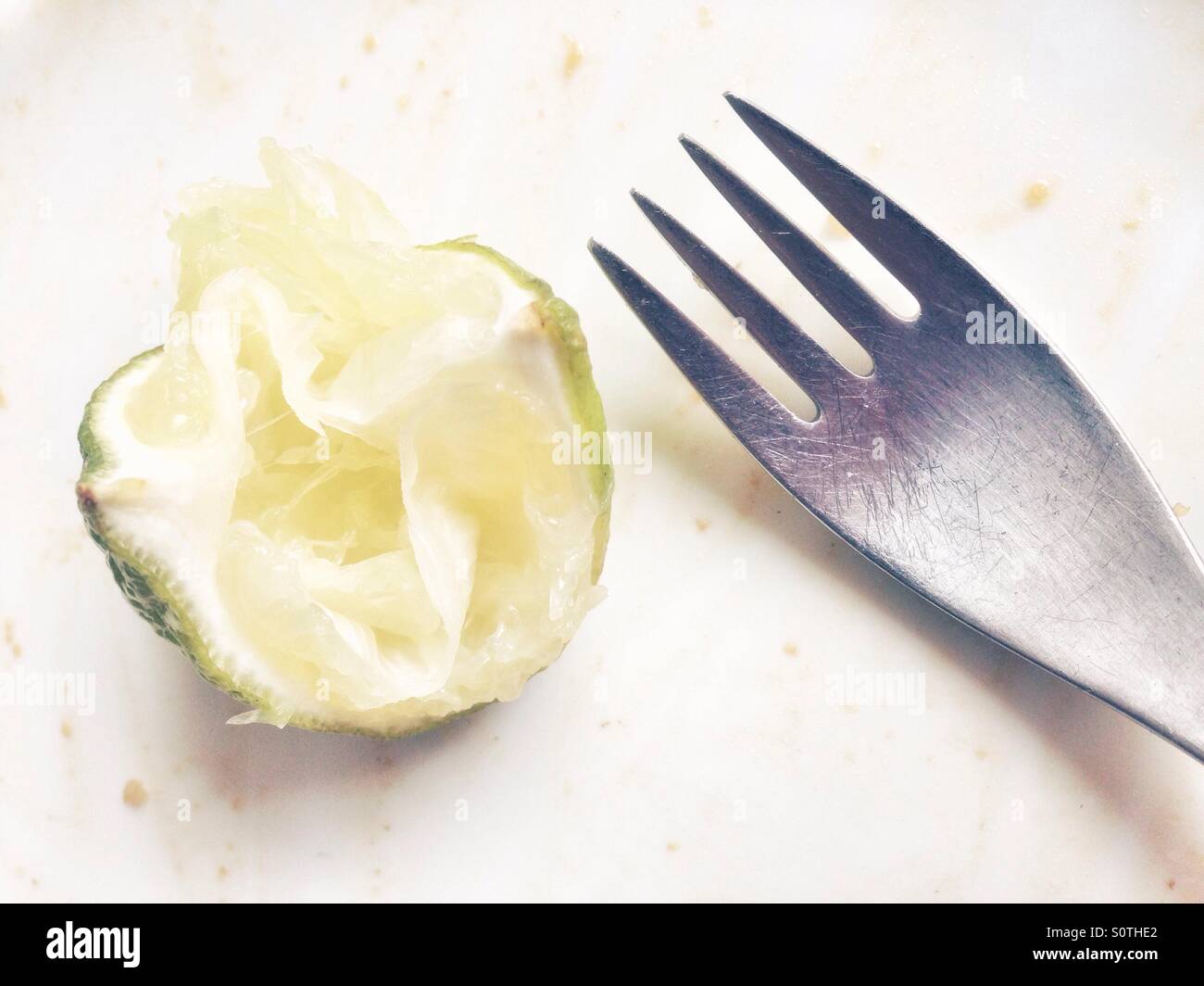 A squeezed lime quarter wedge leftover and a fork on a white plate after a meal - Smartphone Captured Stock Image