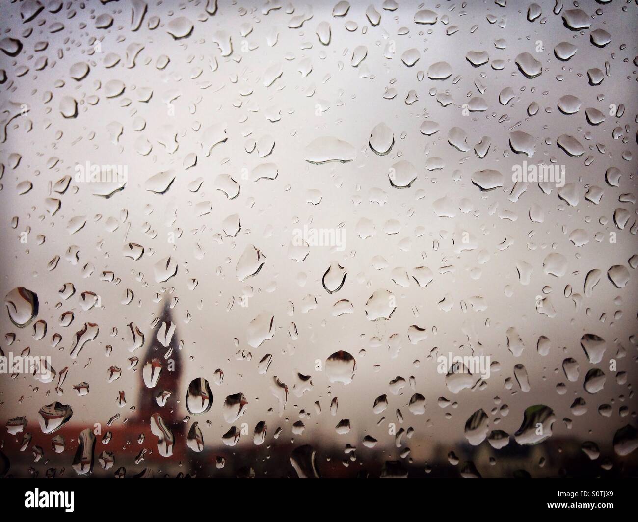 Raindrops on a window with grey sky and the silhouette of a city and a church spire in the background - Smartphone Captured Stock Image