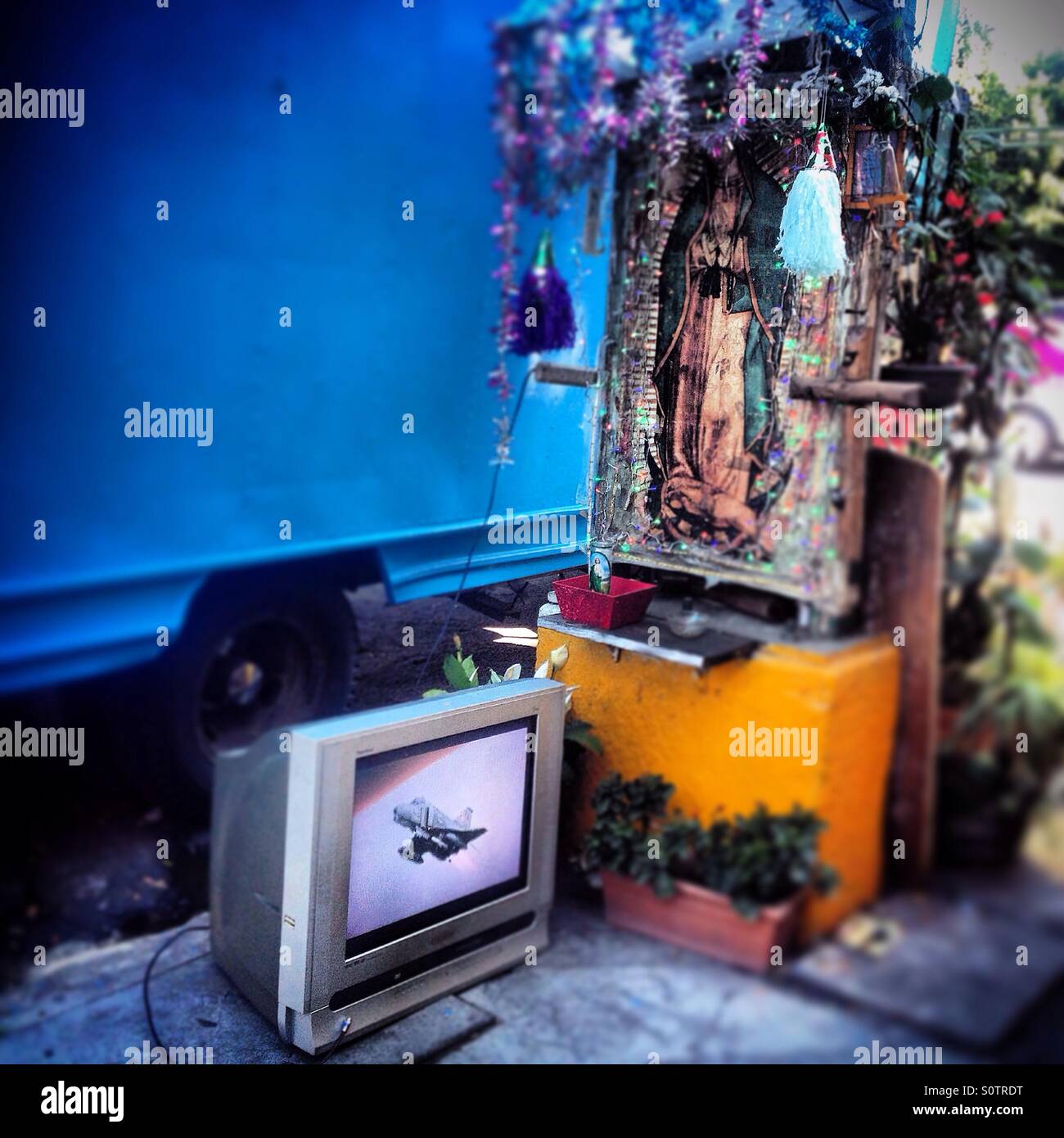 A tv showing a war plane near an altar to Our Lady of Guadalupe, in Mercado de Medellin, Colonia Roma, Mexico City, Mexico Stock Photo