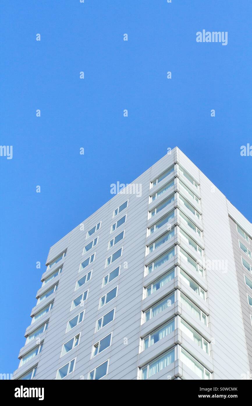 Looking up from the ground to a white, high rise building with uniform Windows and balconies under a bright blue sky. Stock Photo