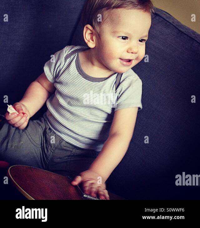 One year old happy toddler with a snack in his hand is touching the guitar Stock Photo