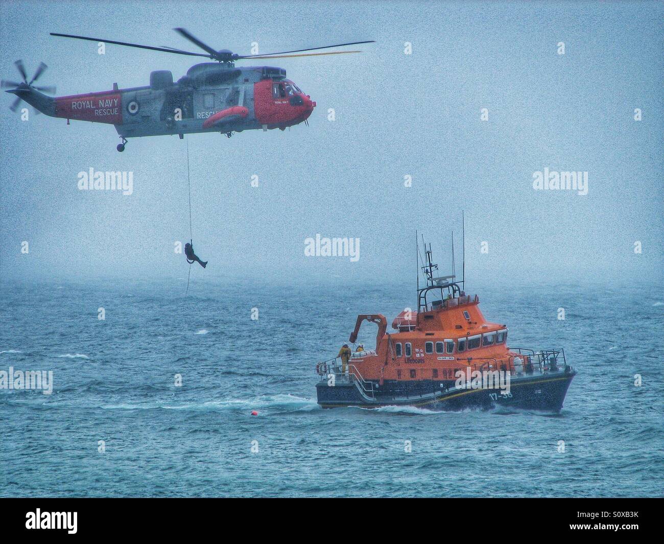 A Royal Navy Air and Sea Rescue helicopter lowering a winch man onto a waiting RNLI lifeboat on a misty ocean. Stock Photo