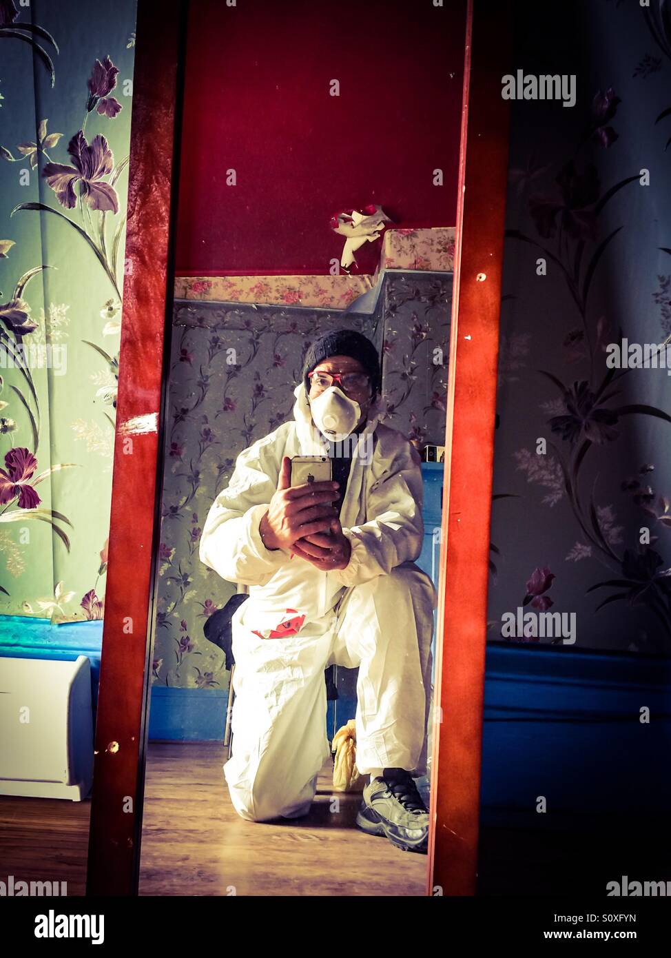Construction worker selfie. Man in overalls wearing a mask kneels down for a photo of his own. Mirror, mirror. A break in routine. A pause. Indoors. Stock Photo