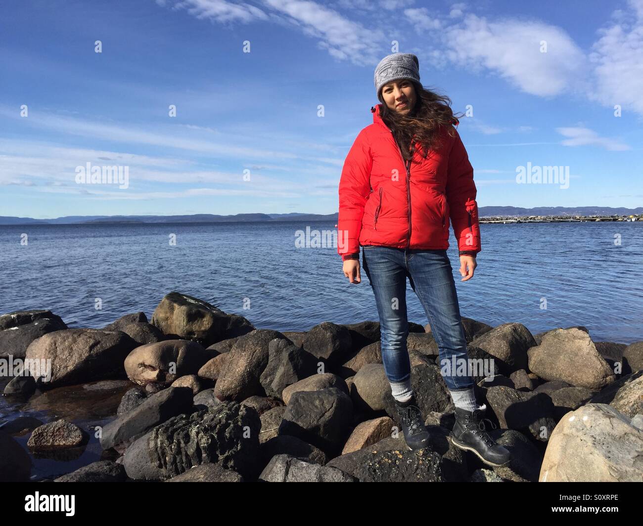 A girl dressed in blue jeans, red jacket and grey hat faces the camera whilst standing on the shoreline on a sunny day in Norway. Stock Photo