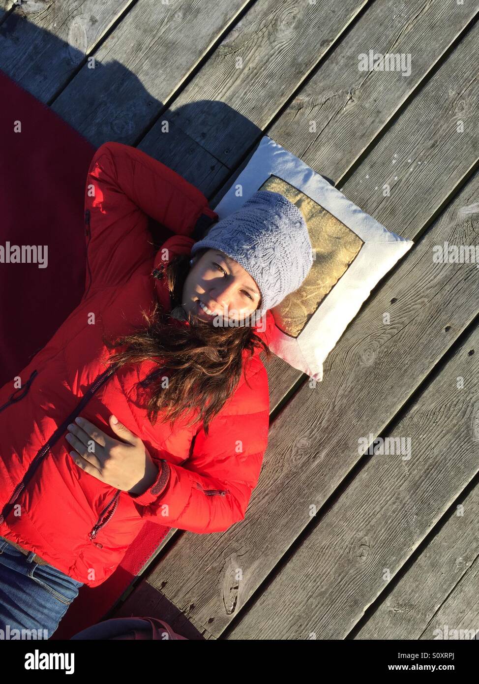 A girl dressed in blue jeans, red jacket and grey hat faces the camera whilst lying on a deck on a sunny day in Norway. Stock Photo