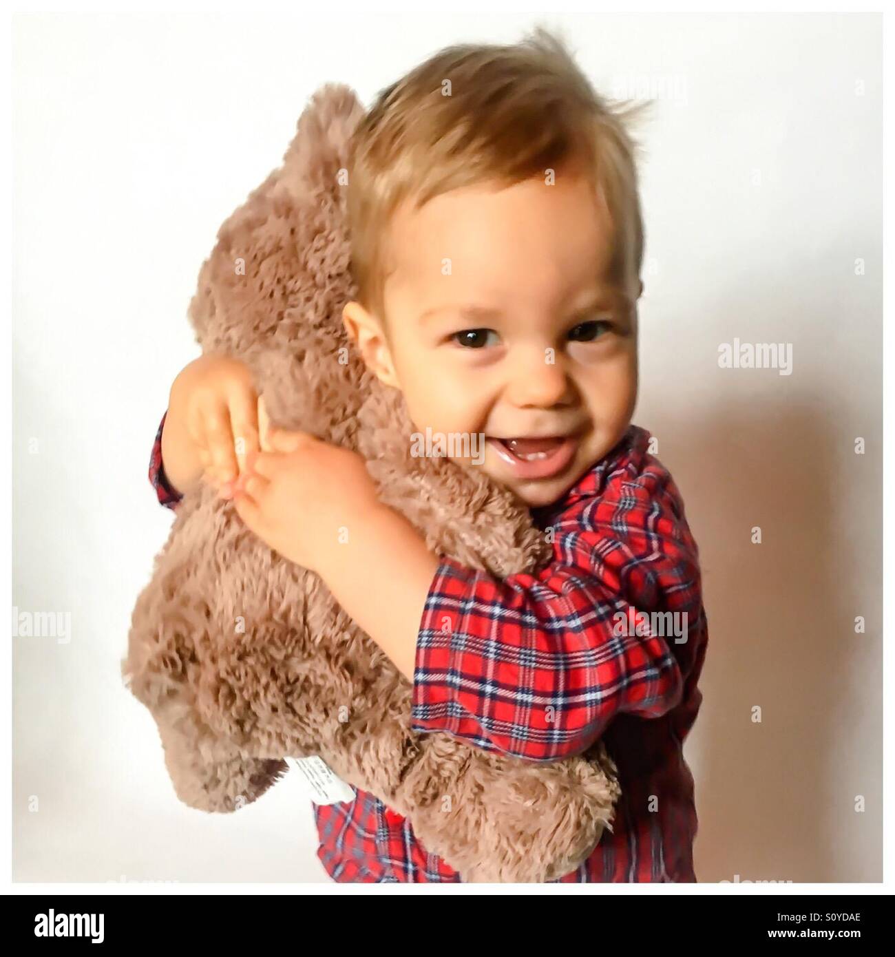 15 months old baby boy wearing red pajamas, smiling and hugging his stuffed teddy bear Stock Photo