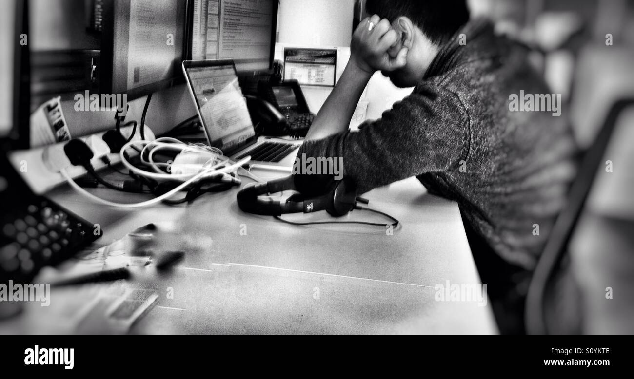 A man rests his head on his hand as he does work on a laptop computer at a desk in an office. - Smartphone Captured Stock Image